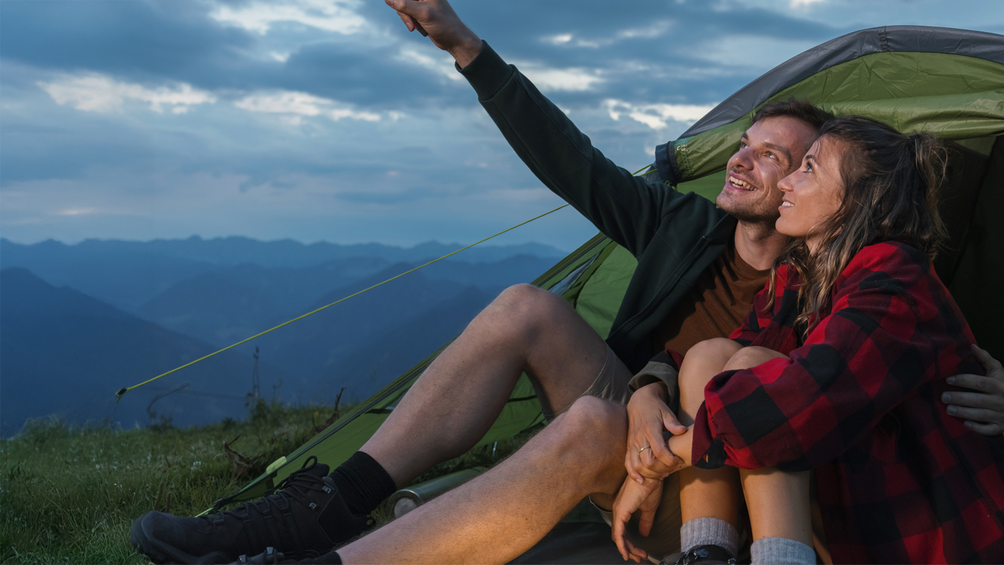 Glückliches Paar sitzt am grünen Zelt auf Berggipfel und zeigt bei Sonnenuntergang zum Himmel