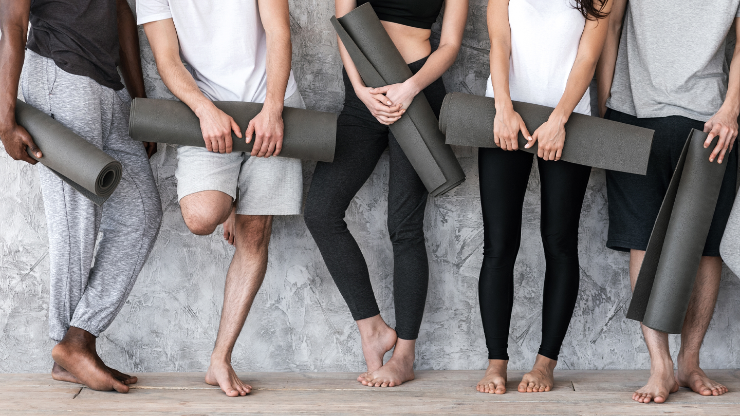 Fünf Personen stehen barfuß mit aufgerollten Yogamatten an einer Wand