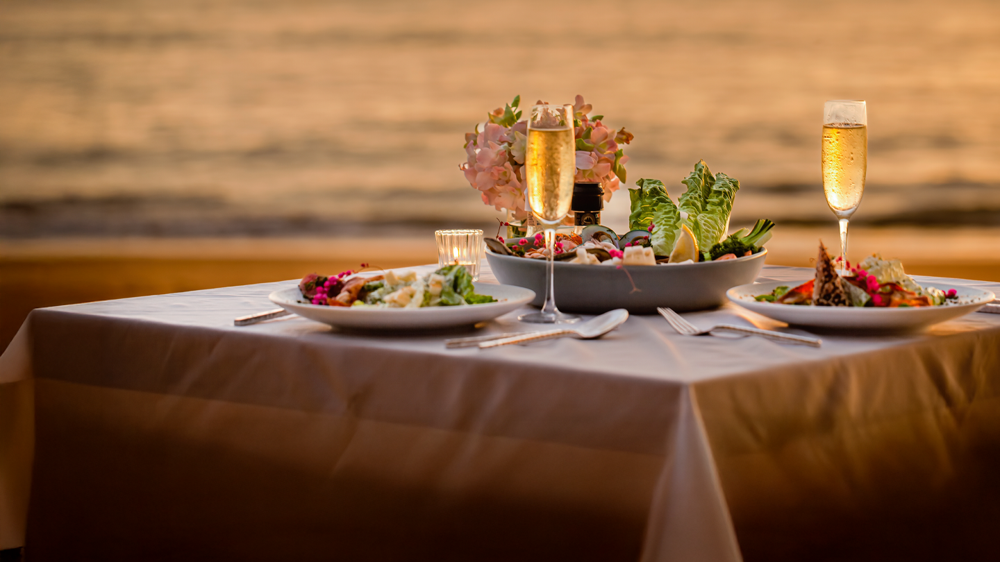 Elegant romantic dinner table set for two on beach at sunset with champagne glasses and gourmet seafood dishes